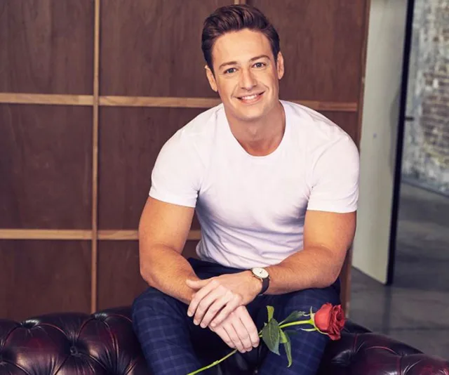 Man in white shirt holding a red rose, seated on a leather sofa, smiling at the camera.