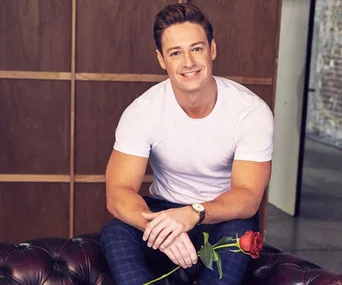 Man in white shirt holding a red rose, seated on a leather sofa, smiling at the camera.