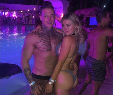 A man and woman pose together by a pool at night with purple lighting and tropical decor in the background.
