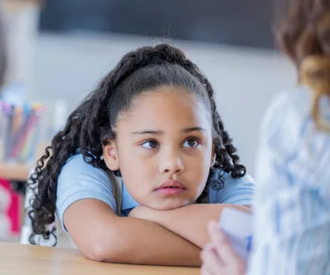 Young girl with curly hair looking up, resting her chin on crossed arms at a classroom desk.