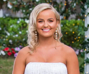 Blonde woman smiling in garden, wearing white dress and pearl earrings.
