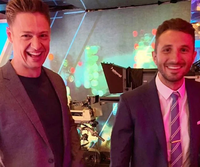 Two men smiling in a brightly lit studio with cameras and colorful background lighting.