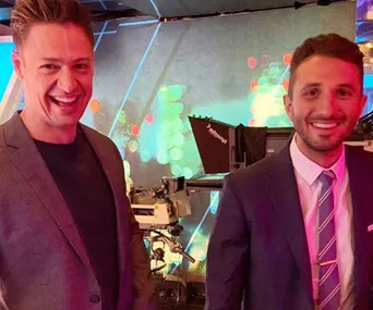 Two men smiling in a brightly lit studio with cameras and colorful background lighting.