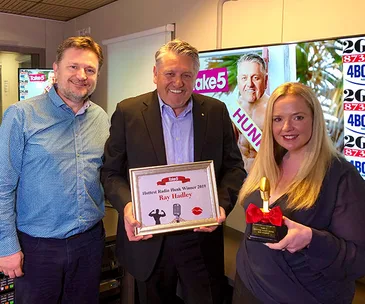 Three people smiling in a studio; one holding a plaque reading "Hottest Radio Hunk Winner 2019 Ray Hadley".