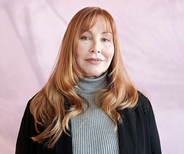 A woman with long red hair, wearing a gray turtleneck and black jacket, standing against a light pink background.