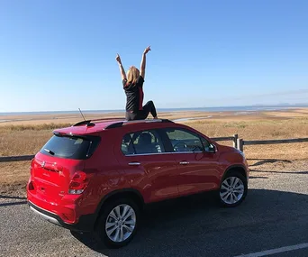 Person sitting on a red SUV with arms raised, overlooking a vast coastal landscape.