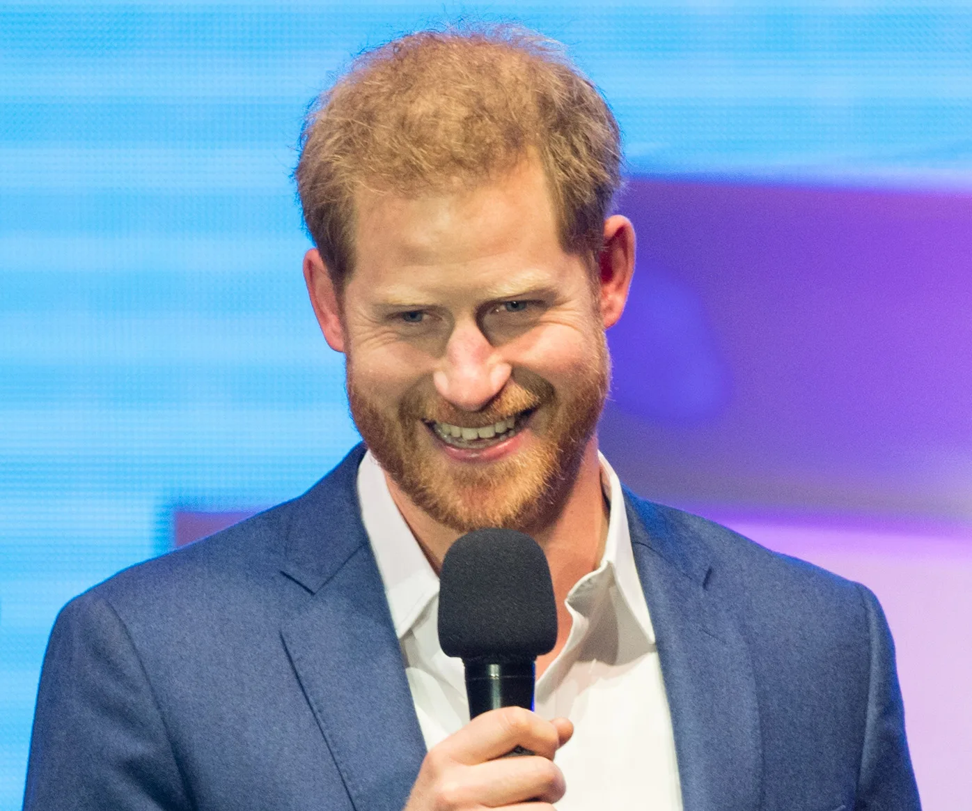 Prince Harry smiling and holding a microphone in a blue suit and white shirt against a colorful background.