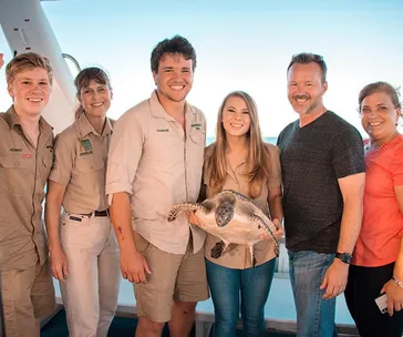 Group of six people smiling on a boat, holding a sea turtle, with ocean in the background.