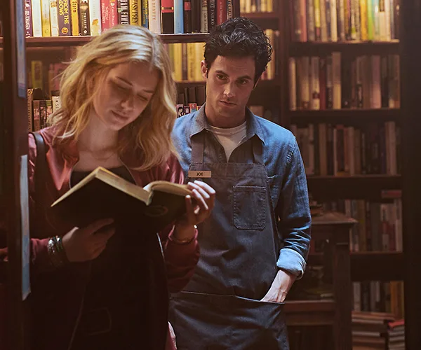 Joe, wearing a name tag, stands by a bookshelf, while a blonde woman reads a book, in a scene from "You" Season 2.