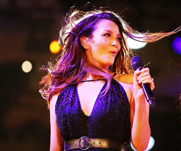 Singer performing on stage, holding a microphone, with hair blowing and wearing a black dress.