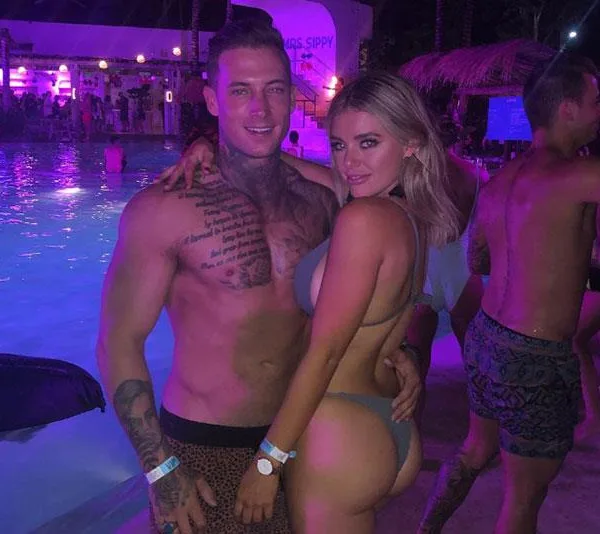 Man and woman in swimwear posing by a lit pool at night, with a crowd and tropical decor in the background.