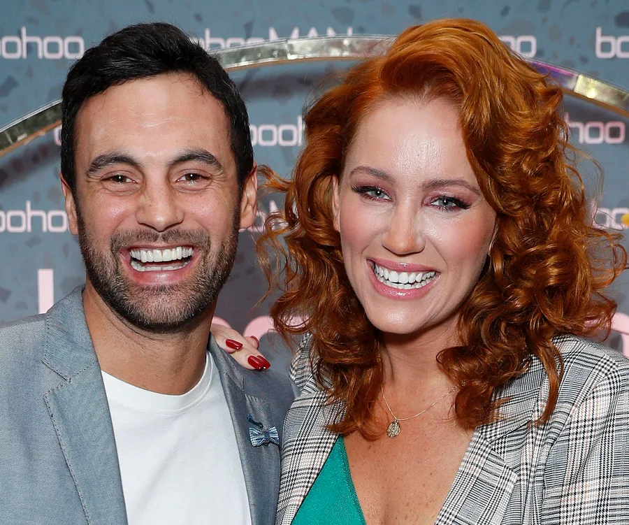 A smiling couple, wearing stylish outfits, stand together at an event with a patterned background.