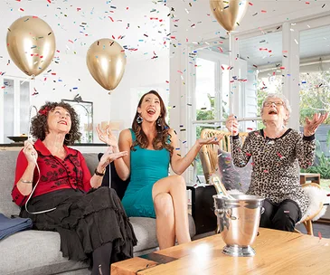 Three women on a couch celebrate with balloons and confetti in a bright living room, with a bucket of champagne on the table.