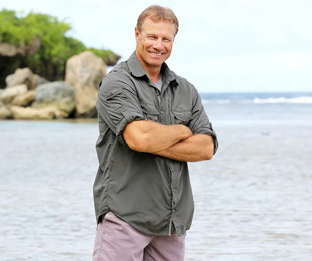 Man smiling by the shoreline with arms crossed, wearing a gray shirt and shorts.