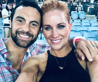 A smiling couple taking a selfie in a stadium with blue seats visible in the background.