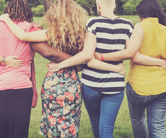 A group of four diverse friends with arms around each other, standing together in a field.