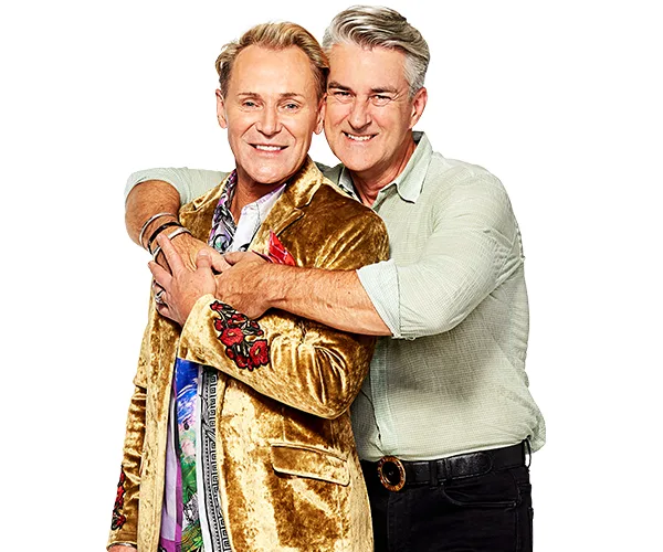 Two men smiling warmly, one in a gold velvet jacket and the other hugging him from behind, against a plain background.