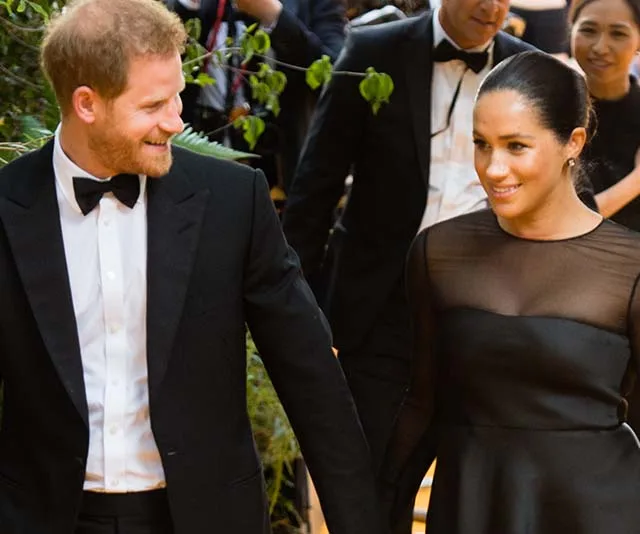 Prince Harry and Meghan Markle smiling and holding hands at a formal event, surrounded by people in evening attire.