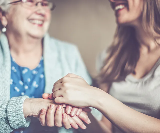Elderly woman and younger woman smiling while holding hands, symbolizing care and support.