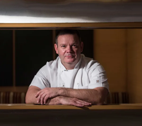 Chef in a white uniform leaning on a countertop, looking directly at the camera, in a kitchen environment.
