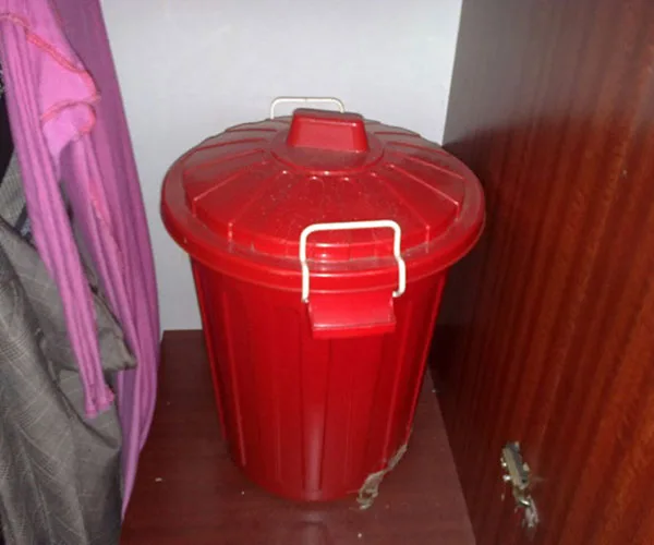 Red plastic trash bin with handles, placed between a wardrobe and a pink garment on a hanger.
