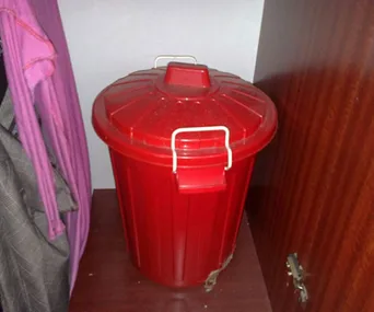 Red plastic trash bin with handles, placed between a wardrobe and a pink garment on a hanger.