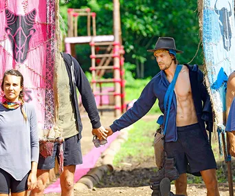 Two contestants from "Survivor" during a challenge, holding hands between pink and blue tribal flags in a jungle setting.