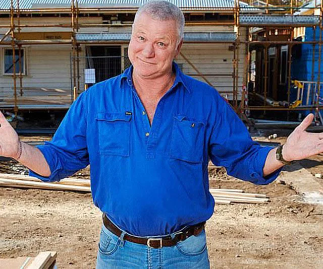 Man in blue shirt gestures with arms open, standing at a construction site with scaffolding and timber.