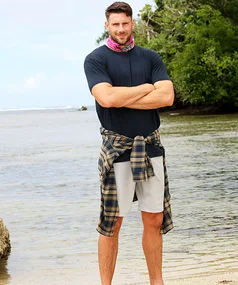 Man standing on a beach with arms crossed, wearing a pink headband, blue shirt, and plaid shirt tied around the waist.