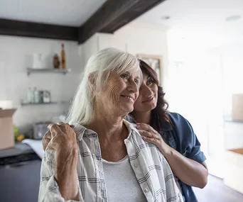 Elderly woman and younger woman embracing in a kitchen, smiling softly.