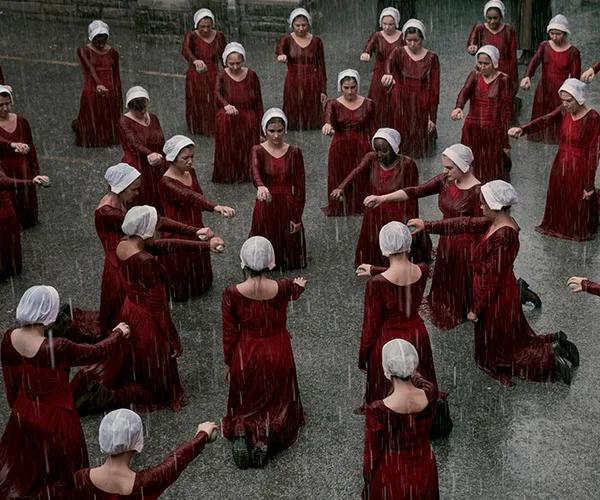 A group of women in red dresses and white bonnets kneeling on wet pavement in the rain, in a dramatic setting.
