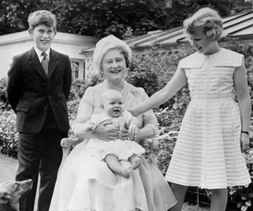 A smiling older woman sits with a baby, flanked by a boy in a suit on the left and a girl in a dress on the right.