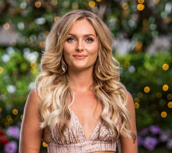 A woman with long blonde hair in a sparkly dress stands outside with fairy lights in the background.