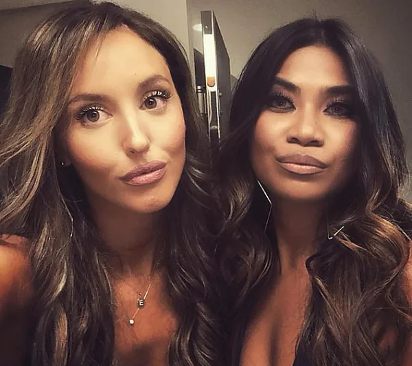 Two women taking a selfie with serious expressions, wearing dark outfits and long hair styled in loose waves.