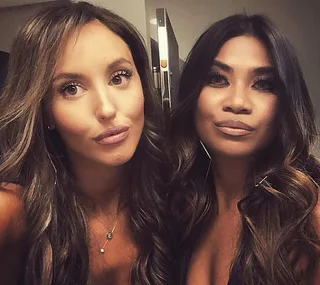 Two women taking a selfie with serious expressions, wearing dark outfits and long hair styled in loose waves.