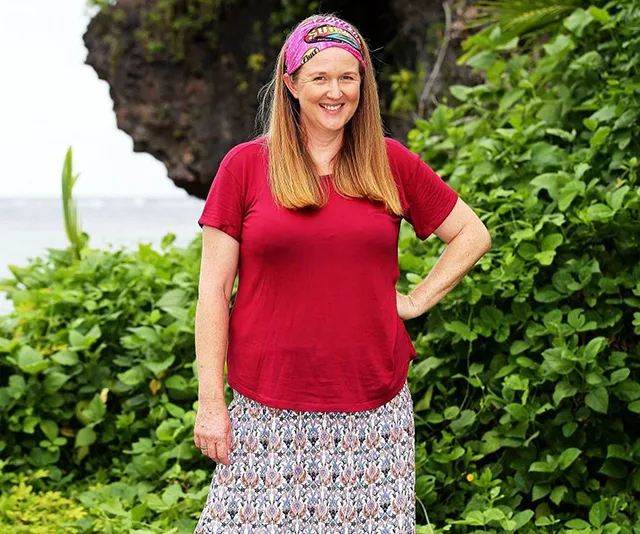 Woman in red shirt and headband stands smiling in lush green setting on "Survivor."