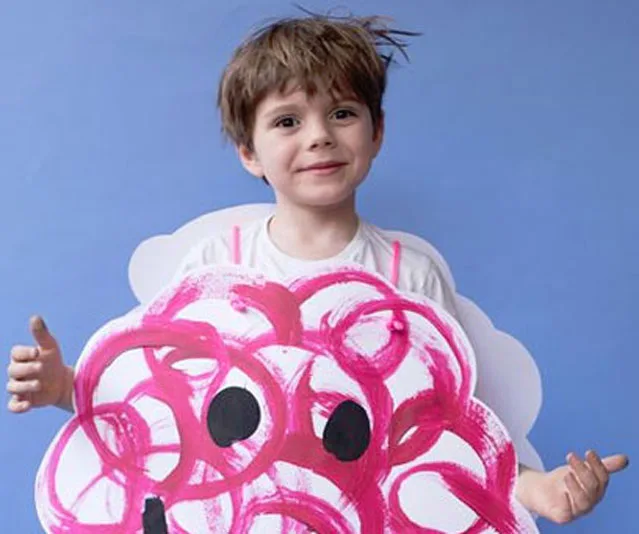 Young child in a painted cloud costume with pink swirls and a happy expression, standing against a blue background.