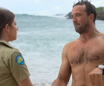 "Two characters in a conversation on a beach from the show 'Seachange'; one wearing a uniform, the other shirtless."