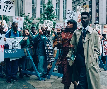Protest scene from "When They See Us" with people holding signs and walking in a crowded street.