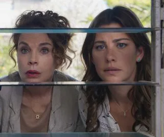 Two women curiously peering through window blinds, sunlight filtering through.