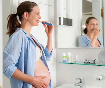 Pregnant woman brushing teeth in bathroom, smiling, wearing a blue shirt and standing by the sink.