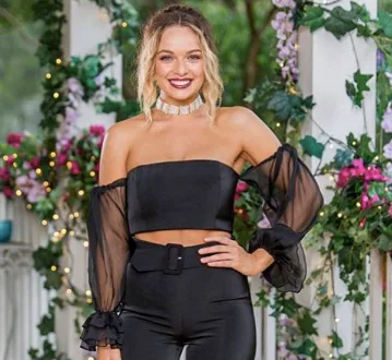 Woman in black off-shoulder top and pants smiling, standing in a floral-decorated setting.