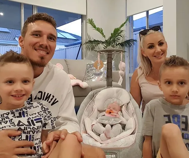 Family posing with baby in car seat, three children and two adults smiling indoors, next to a sofa with a teddy bear.