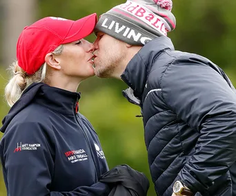A couple shares a kiss outdoors, both wearing hats and jackets, in a natural setting.