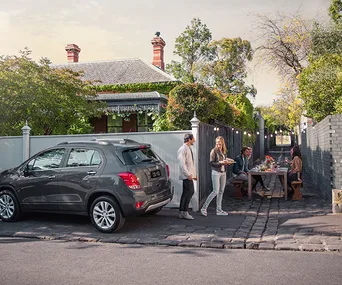 A car parked by a house, with two people walking and others dining outdoors in a garden setting.