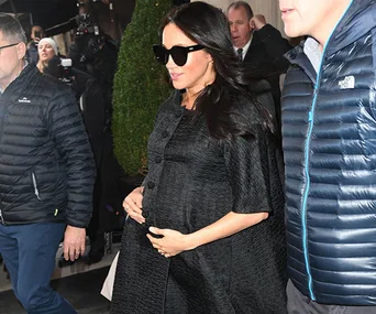 Pregnant woman in a black coat with sunglasses, walking outside surrounded by people in jackets.