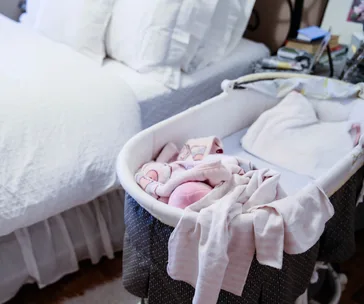 A bassinet with pink baby clothes next to a neatly made bed, in a cozy bedroom setting.