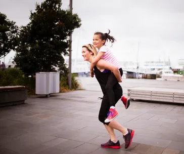 Mother and daughter having fun with piggyback ride in a park near a marina.