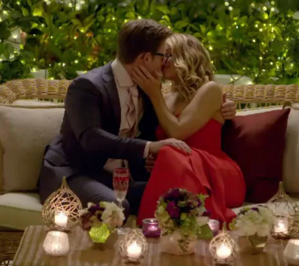 A couple kissing on a candlelit outdoor couch, surrounded by decorative lights and flowers, with greenery in the background.