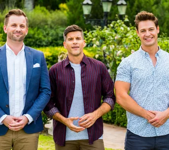 Group of three men smiling outdoors, wearing casual and smart attire, with greenery in the background.